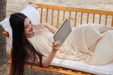 Téléchargez les photos : Jeune femme reposant sur un banc sur une plage de sable fin et regardant un nouvel épisode de sitcom sur tablette - en image libre de droit