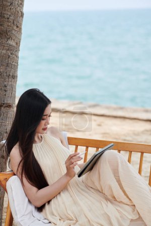 Téléchargez les photos : Jeune femme en robe de soie passer du temps sur la plage et contrat de relecture sur tablette - en image libre de droit