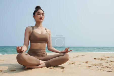 Téléchargez les photos : Jeune femme asiatique méditant en position lotus sur une plage ensoleillée - en image libre de droit