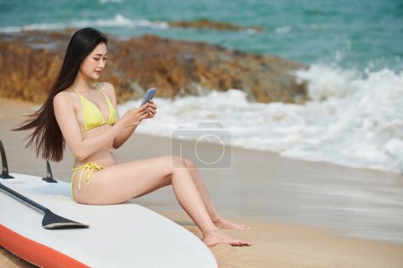 Photo for Joyful young woman checking messages on social media when sitting on supboard on beach - Royalty Free Image