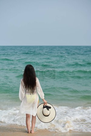 Photo for Young woman standing on sandy beach, taking off hat and enjoying sea waves - Royalty Free Image