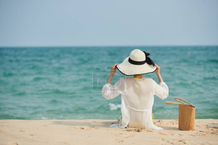 Photo for Young woman sitting on sandy beach in satin shirt and hat and looking at seascape - Royalty Free Image