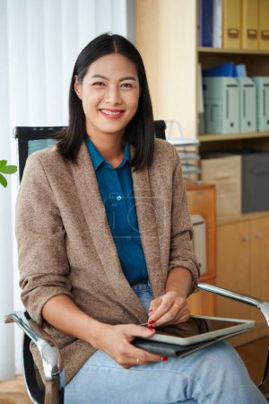 Foto de Retrato vertical de la joven gerente sentada en su silla de oficina con tableta digital y sonriendo a la cámara - Imagen libre de derechos