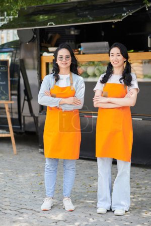 Foto de Dos jóvenes y felices vendedoras asiáticas de comida callejera mirando a la cámara con sonrisas mientras se paran contra un camión en un entorno urbano - Imagen libre de derechos