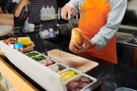 Photo for Close-up of young food vendor in orange apron cutting fresh bun over wooden chopping board while preparing hotdogs for customers - Royalty Free Image