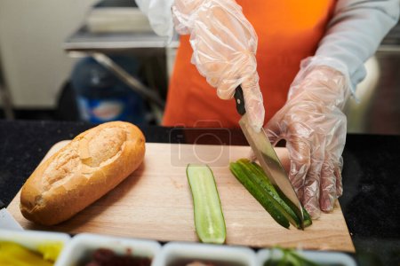 Téléchargez les photos : Mains de femme en gants de protection tranchant du concombre frais sur une planche à découper en bois tout en préparant un hot-dog végétarien dans un camion alimentaire - en image libre de droit