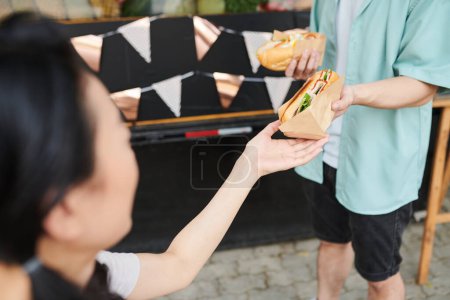 Téléchargez les photos : Gros plan de la jeune femme prenant un hot-dog appétissant frais enveloppé dans du papier de la main de son petit ami en tenue décontractée debout devant - en image libre de droit