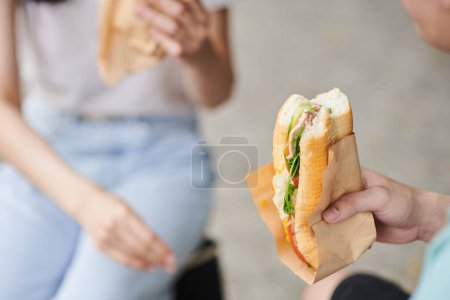 Photo for Close-up of fresh appetizing hotdog with vegetables, bacon and cheese held by hungry young man eating it after buying in street food truck - Royalty Free Image