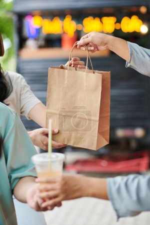 Téléchargez les photos : Focus sur sac en papier avec hot-dogs frais transmis par le vendeur de camion de nourriture de rue aux jeunes consommateurs masculins et féminins après paiement - en image libre de droit