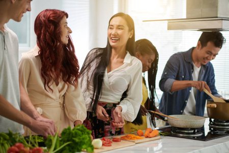 Foto de Feliz joven asiática en ropa de casualwear hablando con sus amigos en la cocina mientras corta verduras frescas y prepara la cena - Imagen libre de derechos