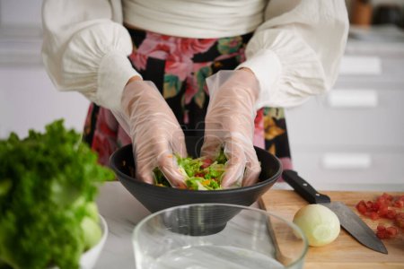 Foto de Primer plano de la mujer joven en guantes mezclando ingredientes de ensalada de verduras en un tazón mientras está de pie junto a la mesa de la cocina y la cena de cocina - Imagen libre de derechos