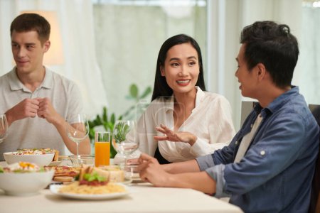 Foto de Joven mujer asiática sonriente hablando con su novio por mesa servida con comida y bebidas caseras apetitosas para la cena o fiesta en casa - Imagen libre de derechos