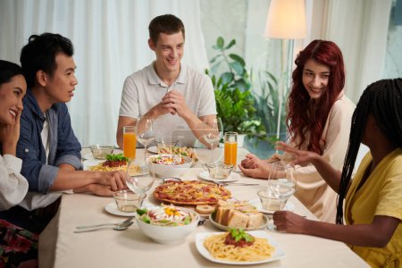 Foto de Grupo de jóvenes interculturales en ropa de casualwear inteligente charlando por la mesa servida y disfrutando de la cena que consiste en comida casera - Imagen libre de derechos