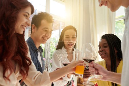 Téléchargez les photos : Joyeux jeunes amis interculturels claquant avec leurs boissons à la fête à la maison devant la caméra tout en profitant de la célébration et en s'amusant - en image libre de droit