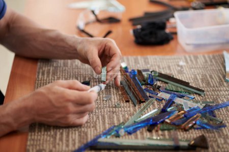 Téléchargez les photos : Gros plan de l'artisan assis sur son lieu de travail et faisant de l'artisanat à partir de verre coloré - en image libre de droit