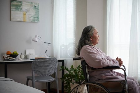Téléchargez les photos : Malade femme âgée assise en fauteuil roulant et regardant par la fenêtre - en image libre de droit