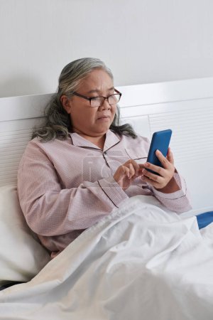Foto de Mujer mayor grave sentada en la cama y leyendo notificaciones y mensajes de texto de amigos - Imagen libre de derechos