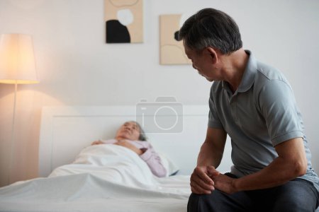 Photo for Senior man sitting on bed of sick wife sleeping in bed - Royalty Free Image