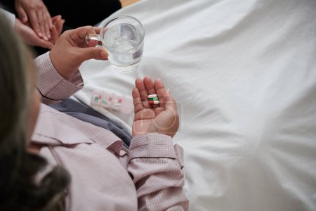 Téléchargez les photos : Malade femme âgée prenant des pilules et de boire une tasse d'eau lorsque vous êtes assis dans le lit - en image libre de droit