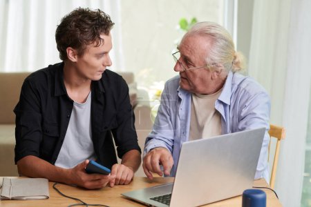 Foto de Hombre joven visitando abuelo para mostrarle cómo utilizar el ordenador portátil para pagar facturas en línea - Imagen libre de derechos