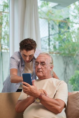 Foto de Hombre mayor pidiendo nieto para ayudarle con la aplicación móvil en el teléfono inteligente - Imagen libre de derechos