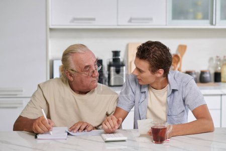 Téléchargez les photos : Jeune homme aidant grand-père à planifier le budget quand ils sont assis au comptoir de la cuisine - en image libre de droit