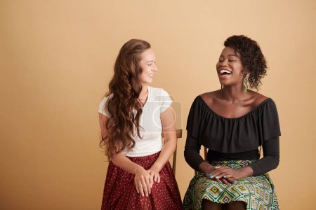 Téléchargez les photos : Jeunes amies multiethniques riant et plaisantant sur des chaises contre un fond beige - en image libre de droit
