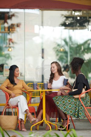 Foto de Grupo de mujeres jóvenes en ropa de verano brillante charlando en la mesa en la cafetería al aire libre - Imagen libre de derechos