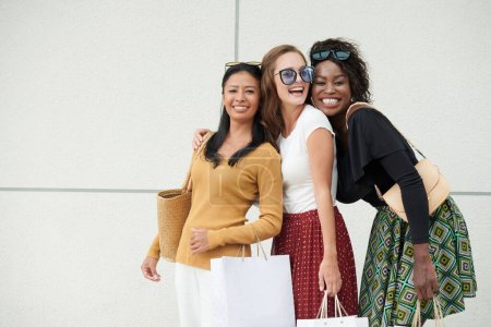 Photo for Portrait of multi-ethnic female friends standing at wall with shopping bags in hands and looking at camera - Royalty Free Image