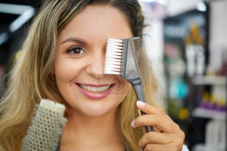 Foto de Cara de estilista sonriente cubriendo un ojo con peine de dientes anchos de plástico para teñir el cabello y mirando a la cámara - Imagen libre de derechos