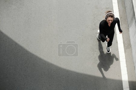 Photo for Young female sportswoman listening to music in headphones when jogging on road in the morning, view from above - Royalty Free Image