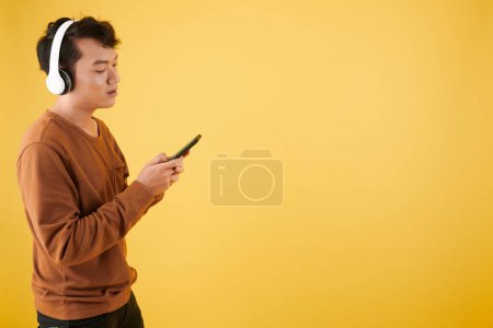 Foto de Hombre joven en auriculares comprobar la lista de reproducción en el teléfono inteligente y elegir qué escuchar - Imagen libre de derechos