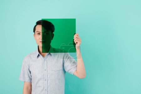 Téléchargez les photos : Jeune homme couvrant la moitié du visage avec un filtre photo en feuille de plastique vert transparent - en image libre de droit