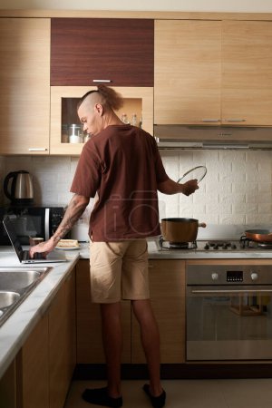 Photo for Young man in loungewear cooking soup for dinner and checking e-mails and notifications on laptop - Royalty Free Image