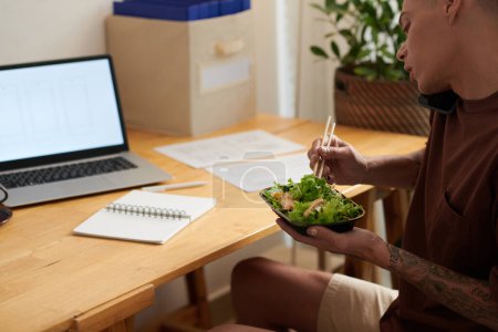 Téléchargez les photos : Jeune homme assis au bureau à la maison, mangeant de la salade de poulet et parlant au téléphone avec un collègue - en image libre de droit