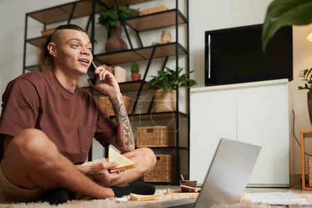 Téléchargez les photos : Jeune homme positif mangeant un sandwich et parlant au téléphone assis sur le sol devant un ordinateur portable ouvert - en image libre de droit