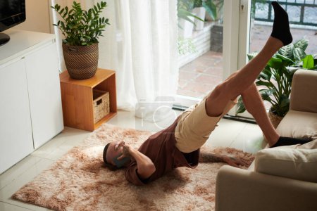 Photo for Young man talking on phone, leaning on couch when doing bridge pose at home - Royalty Free Image