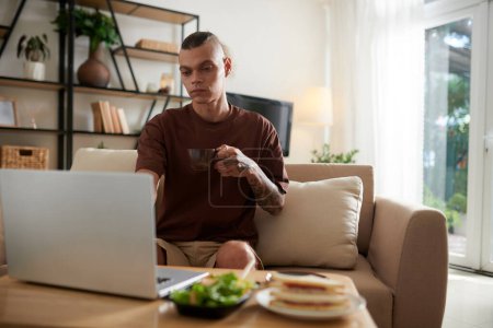 Téléchargez les photos : Un jeune homme sérieux qui boit une tasse de café et prend son petit déjeuner en regardant un webinaire ou un spectacle sur ordinateur portable - en image libre de droit