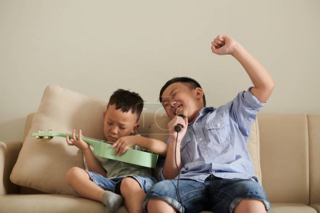 Photo for Emotional boy singing into microphone when his little brother playing ukulele at home - Royalty Free Image