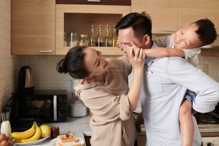 Foto de Esposa e hijo pequeño cubriendo los ojos de padre feliz cuando están de pie en la cocina por la mañana - Imagen libre de derechos