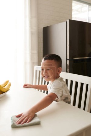 Téléchargez les photos : Petit garçon souriant essuyant table de cuisine avec chiffon doux après le petit déjeuner - en image libre de droit