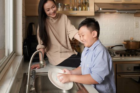 Foto de Feliz madre sentada en el mostrador de la cocina y mirando a su hijo lavar los platos después de cenar - Imagen libre de derechos