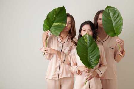 Foto de Tres mujeres jóvenes en pijama de seda rosa claro sosteniendo grandes hojas verdes - Imagen libre de derechos