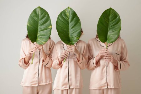 Photo for Three young women in light pink silk pajamas holding faces behind big green leaves - Royalty Free Image