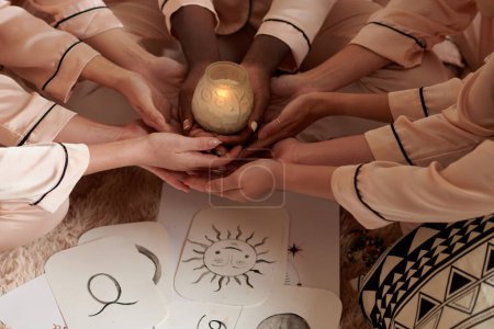 Foto de Manos de amigos sosteniendo vela encendida sobre tarjetas de papel con signos del zodiaco dibujados a mano - Imagen libre de derechos