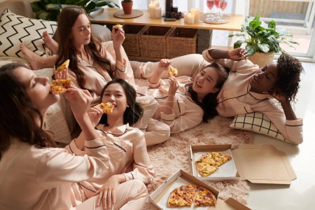 Téléchargez les photos : Joyeux jeune femme en pyjama passer du temps ensemble, bavarder, manger de la pizza et s'amuser - en image libre de droit