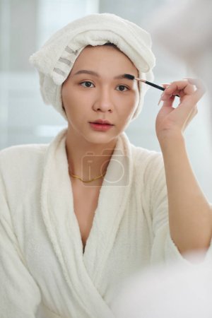 Téléchargez les photos : Transgenre femme brossant les sourcils lors de la préparation après la douche du matin - en image libre de droit