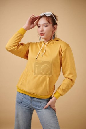 Téléchargez les photos : Portrait de jeune femme transgenre en sweat à capuche jaune et jean posant sur fond beige - en image libre de droit