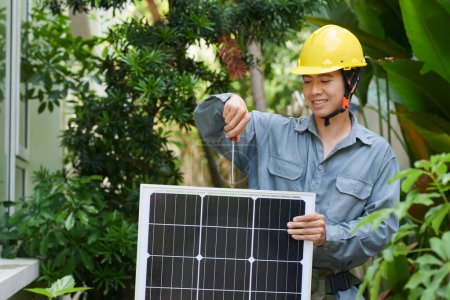 Téléchargez les photos : Portrait d'un installateur souriant de panneaux solaires portant un casque rigide vissant un panneau solaire - en image libre de droit