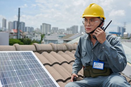 Photo for Contractor standing on roof next to solar panel and talking using walkie-talkie - Royalty Free Image